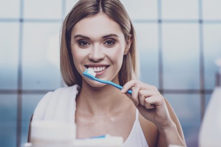 Whitening. Pleasant young woman with attractive appearance and a towel thrown over her shoulder cleaning teeth while looking at camera with cheer on her face