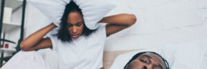 unhappy african american woman covering head with pillow while sitting in bed near snoring husband