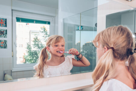 close up portrait of beautiful charming girl with blond braided hair cleaning her teeth in the morning in front of the mirror